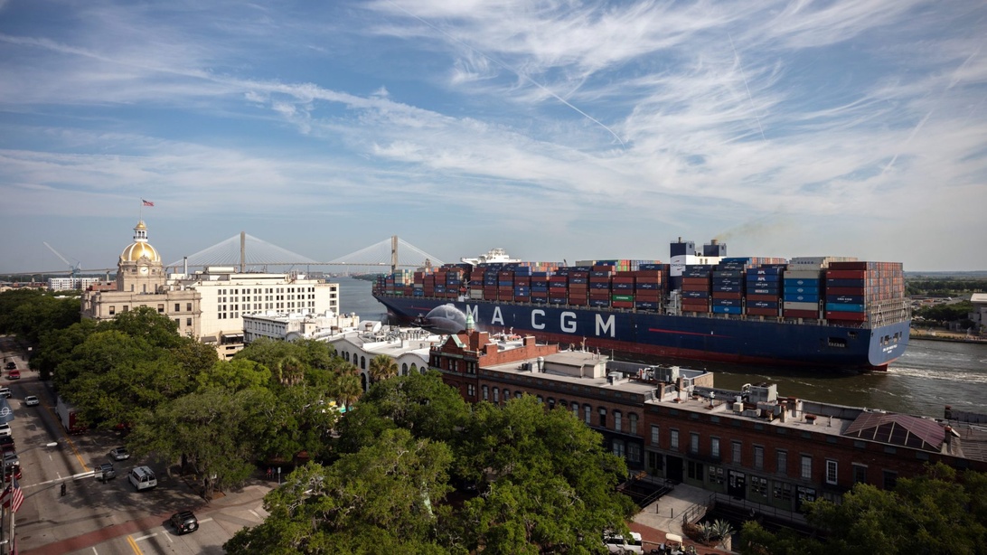 En flygvy över ett massivt CMA CGM containerfartyg fullastat med containrar, navigerande nära ett stadsområde med historiska byggnader, vilket illustrerar global handel.