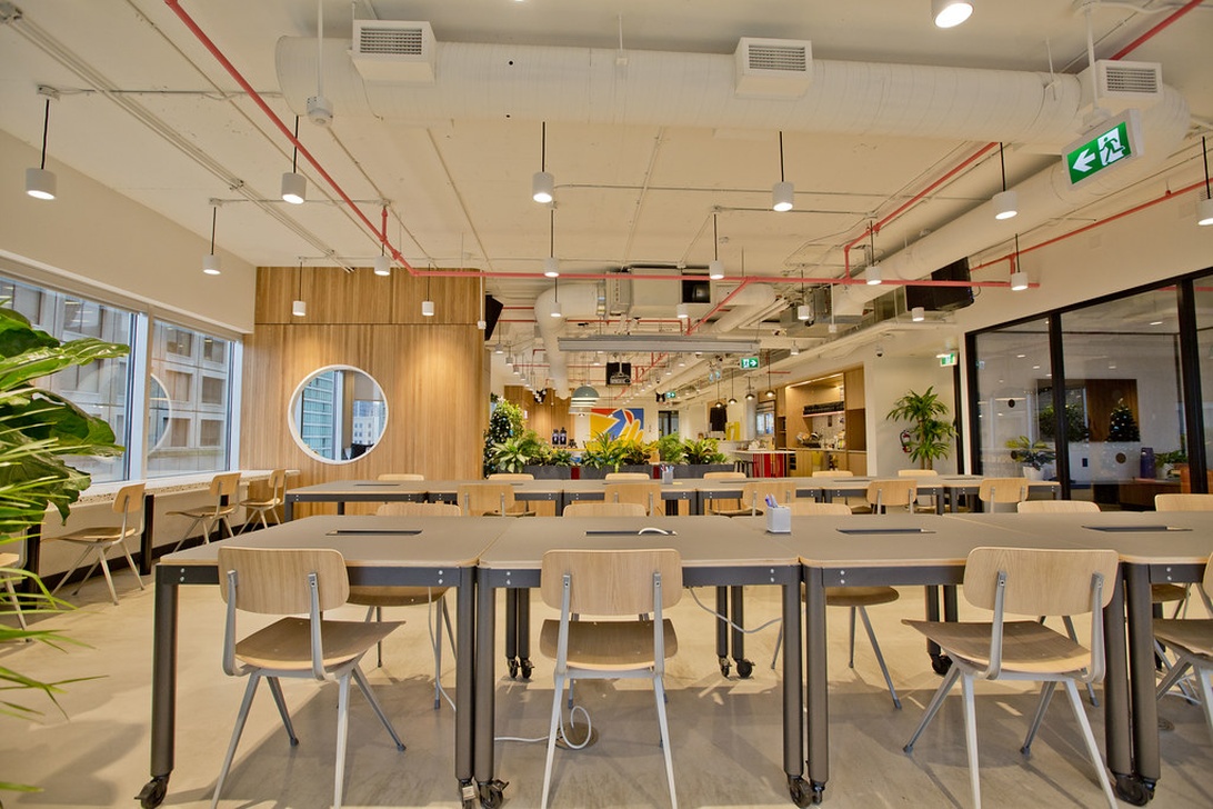 En ljus och modern coworking-yta med gemensamma träbord, ljusa stolar och synliga takinstallationer. Stora fönster ger rikligt med dagsljus och gröna växter är integrerade i miljön.