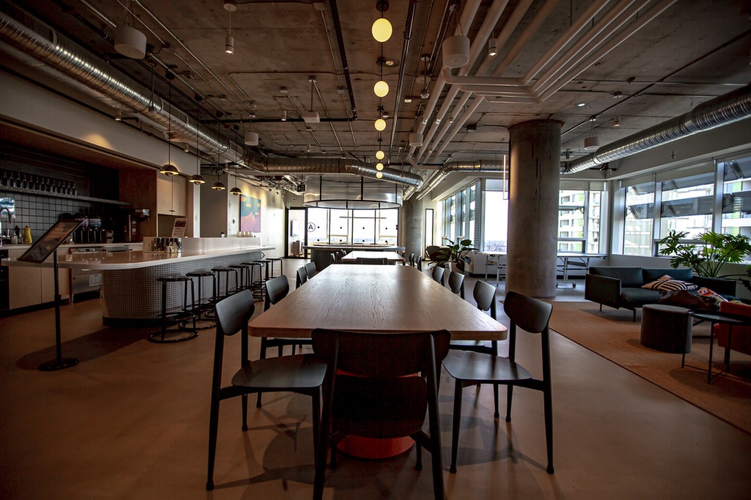 En coworking-yta i industriell stil med synliga takinstallationer, pendelbelysning och ett stort gemensamt träbord. Till vänster finns en bar/kafé-del och i bakgrunden syns loungemöbler.