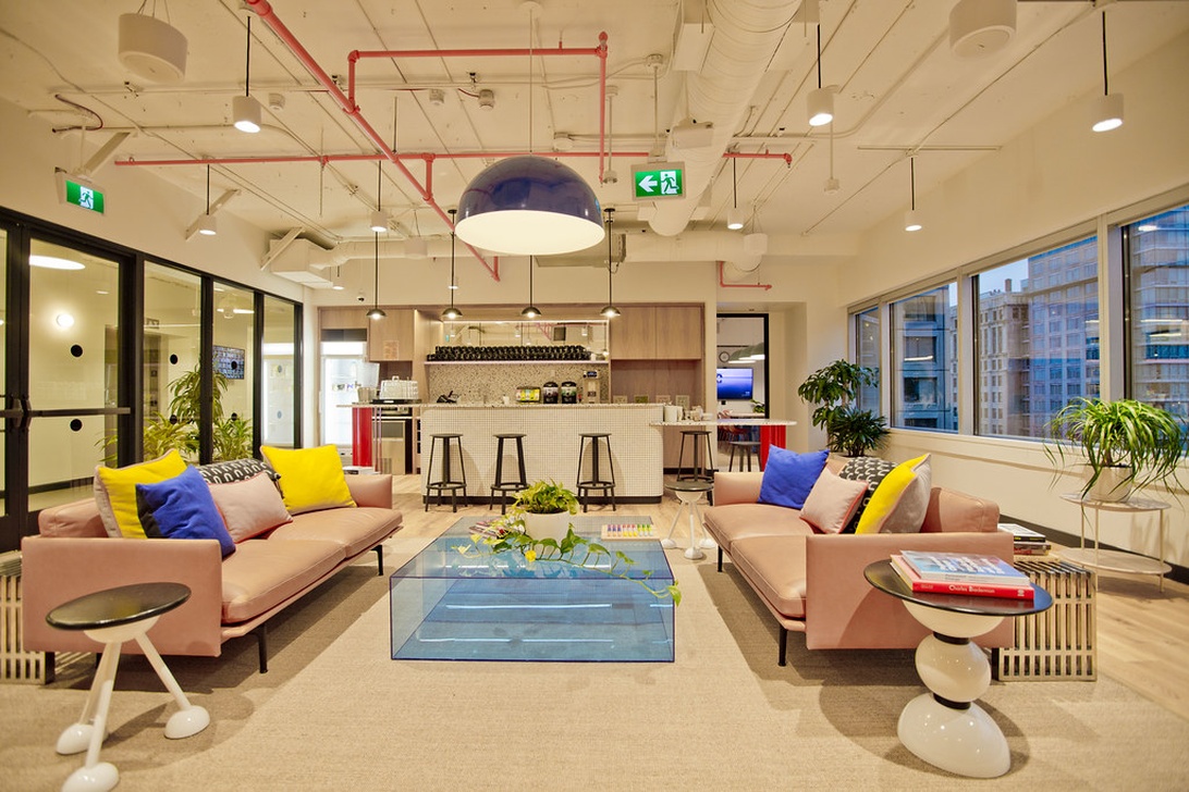 En livfull coworking-lounge med rosa soffor, färgglada kuddar, ett soffbord i glas och en liten förfriskningsbar i bakgrunden. Utrymmet har moderna designelement och stora fönster.