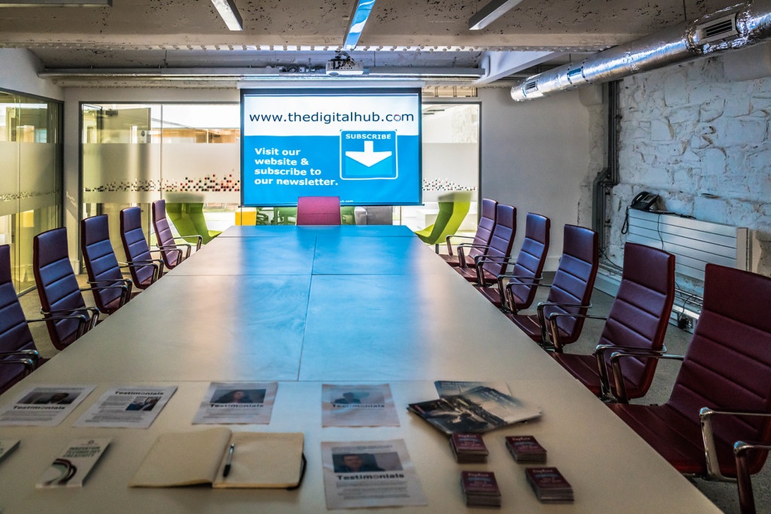Ett modernt konferensrum med ett avlångt bord omgivet av lila och gröna kontorsstolar. Rummet har industriella designelement med synliga rör och texturerat tak. En stor projektionsskärm som visar en webbadress dominerar ena väggen.