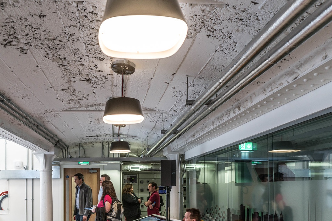 En korridor eller ett gemensamhetsutrymme i en modern kontorsbyggnad med industriell estetik, exponerat tak, pendellampor och glasväggar. Flera personer samtalar i bakgrunden.
